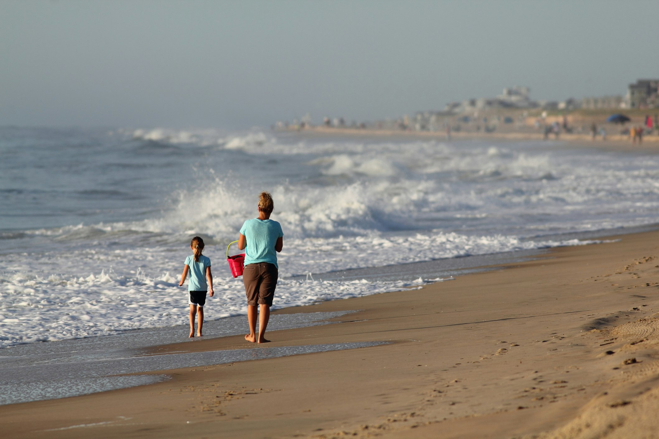 A mother and her young daughter walk along a beach in the morning. The waves are high and the mother is carrying a red bucket
