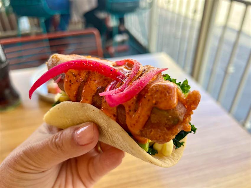 A hand holding a crispy avocado taco with pickled red onion, chipotle aioli, and marinated kale from El Granjero in LA