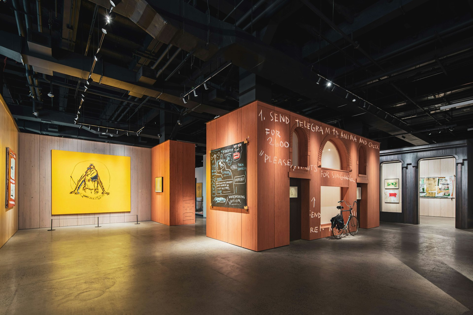 An installation view of the Jean-Michel Basquiat: King Pleasure exhibit in the Starrett-Lehigh Building in West Chelsea, New York. A yellow piece hangs on the back wall and to the right, a small stand-alone structure resembling a brownstone building, with a bike parked in front. 