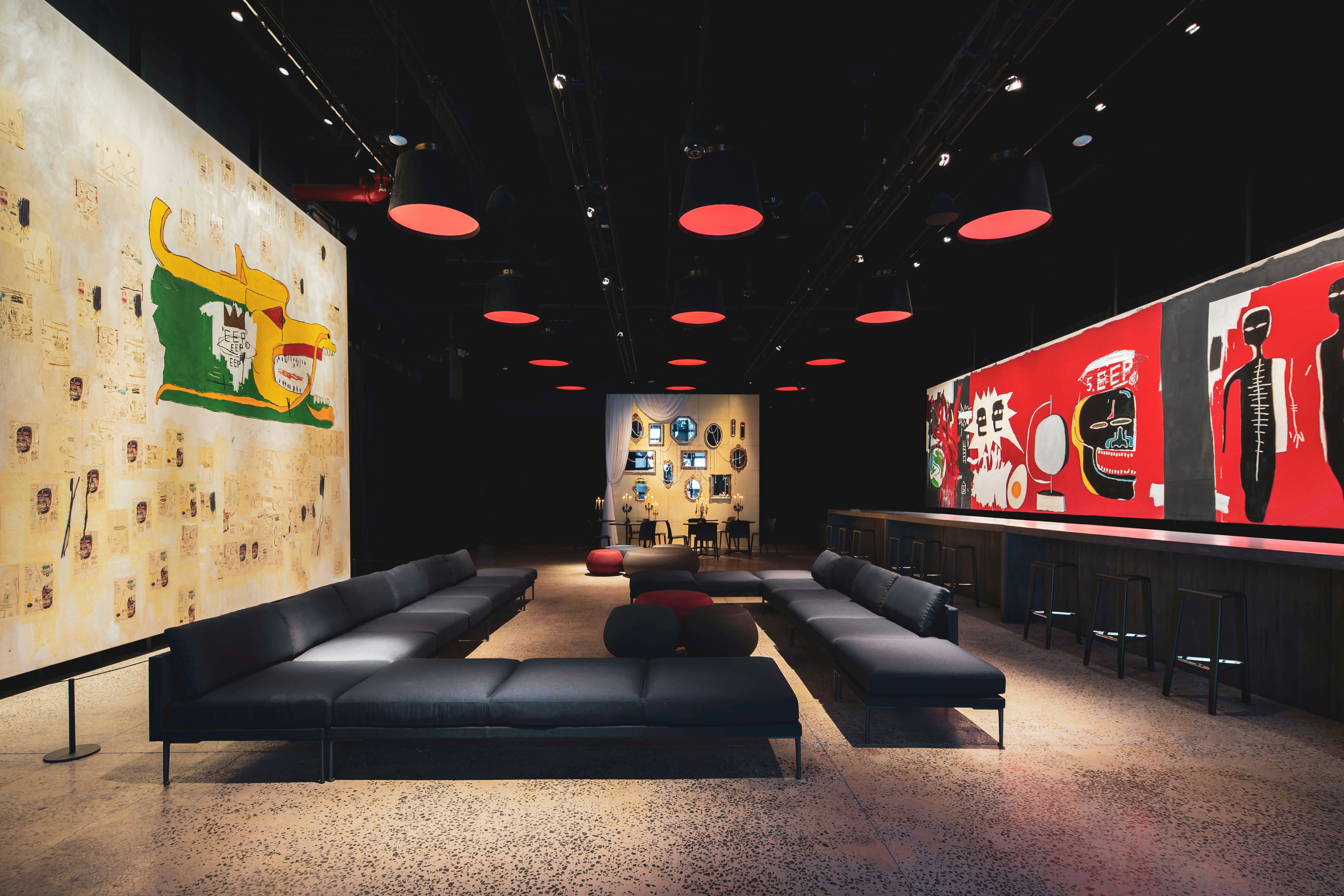 Recreation of the Michael Todd VIP Room of NYC’s iconic Palladium nightclub, for which Basquiat created two paintings. Round red pendant lamps overhead, a bar with high stools and a large red piece on the right, a huge white piece on the left, and low-slung black sofas in the middle