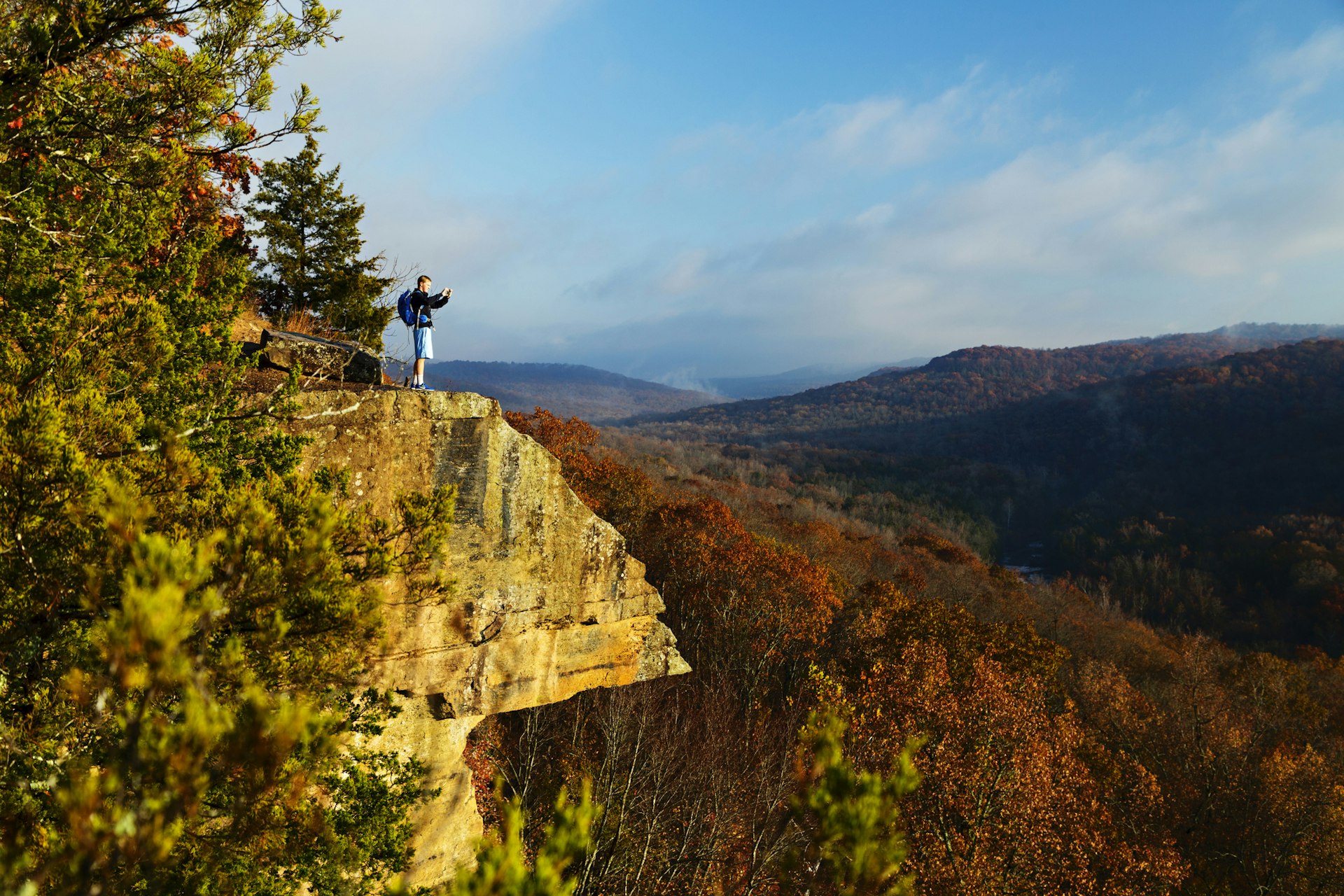 A solo hiker stands on a rocky outcrop taking a photo of the landscape in Devil's Den State Park, Arkansas