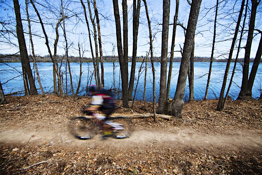 A mountain biker is blurred as they speed along a tree-lined woodland path near a lake in the Ozarks