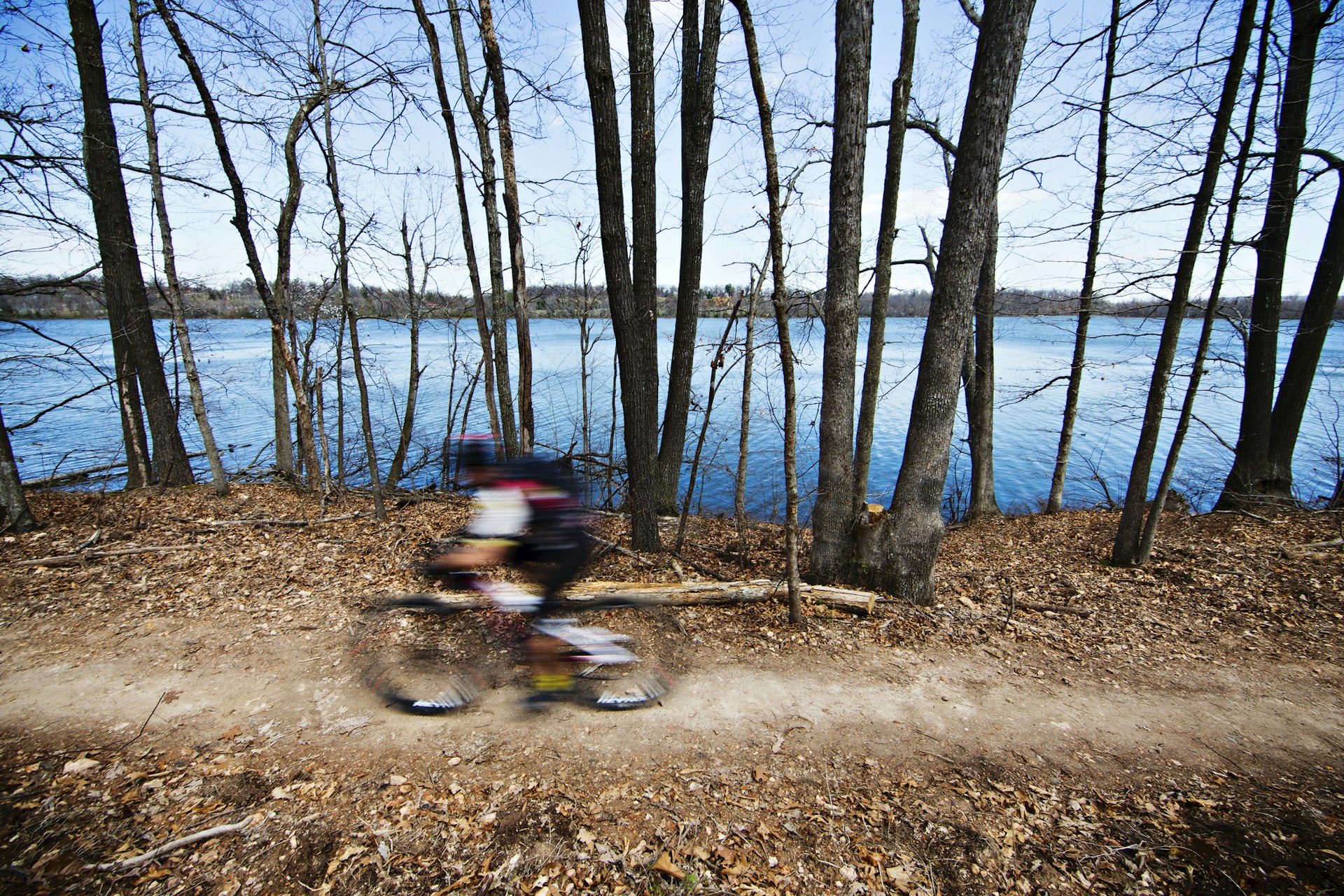 A mountain biker is blurred as they speed along a tree-lined woodland path near a lake in the Ozarks