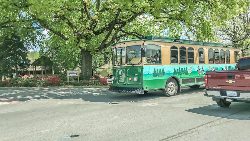 A green and brown Fun Time Trolley, painted with cartoon bears and trees on the side, under a tree by a trolley stop in Pigeon Forge, Tennessee, 
