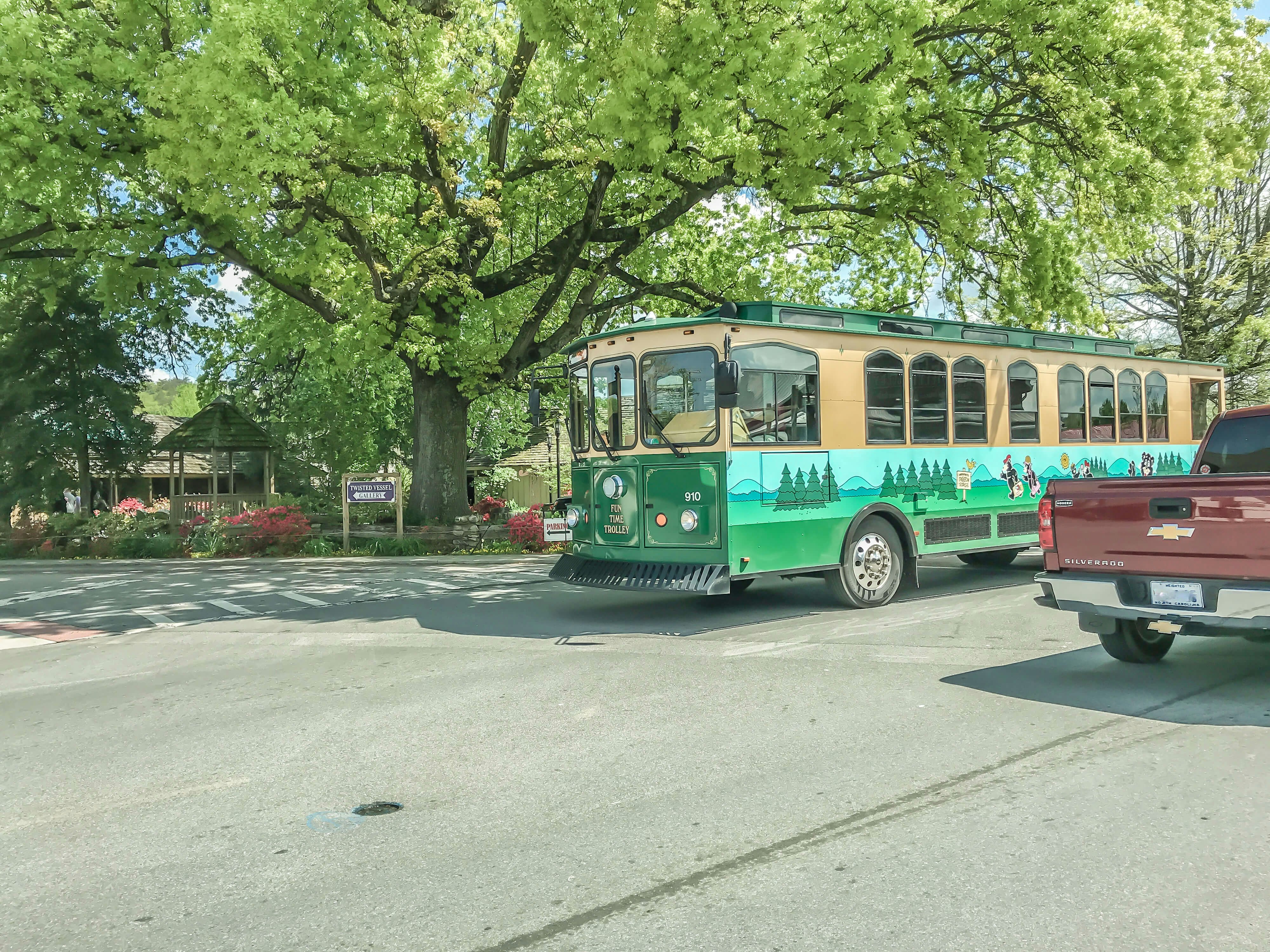 A green and brown Fun Time Trolley, painted with cartoon bears and trees on the side, under a tree by a trolley stop in Pigeon Forge, Tennessee, 