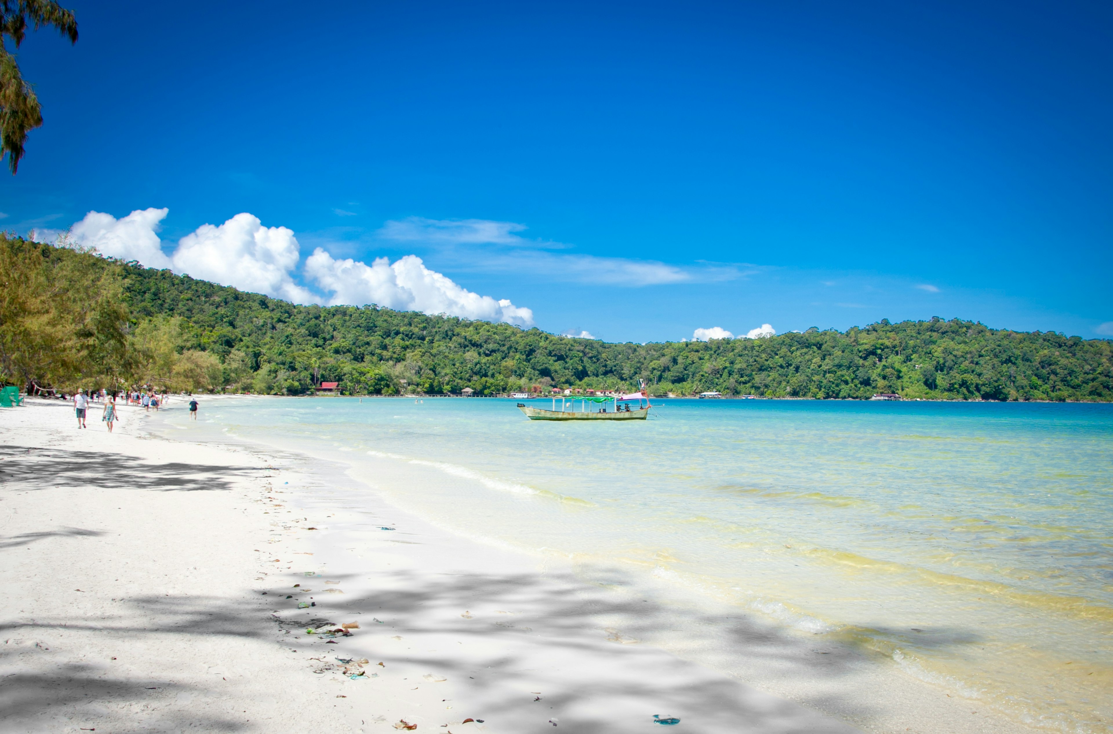 A wide white-sand beach with people and a small boat in turquoise water and a low green mountain ridge in the distance