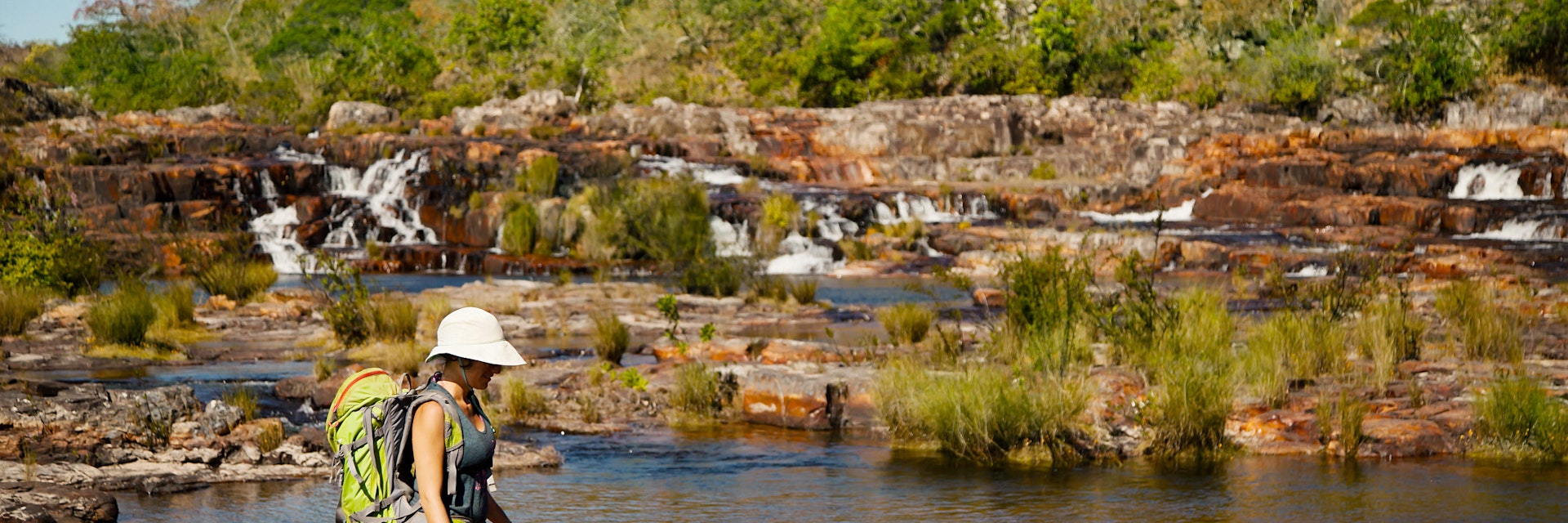 A woman hiking over rocks with a waterfall in the background at Chapada dos Veadeiros National Park.