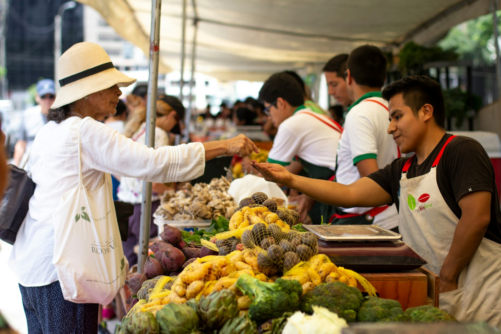 A woman in a white shirt and hat hands money to a vegetable seller at a market in Milaflores, Lima, Peru