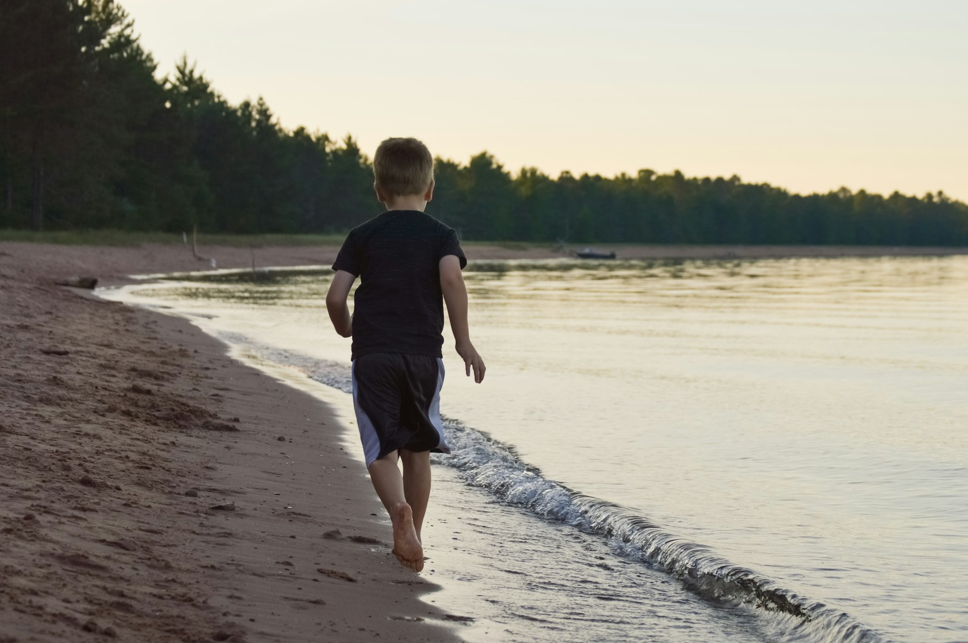 School aged boy runs on a deserted sandy beach surrounded by evergreen forest with a boat beached in the distance under yellow evening sky, Northern Wisconsin, Great Lakes, USA