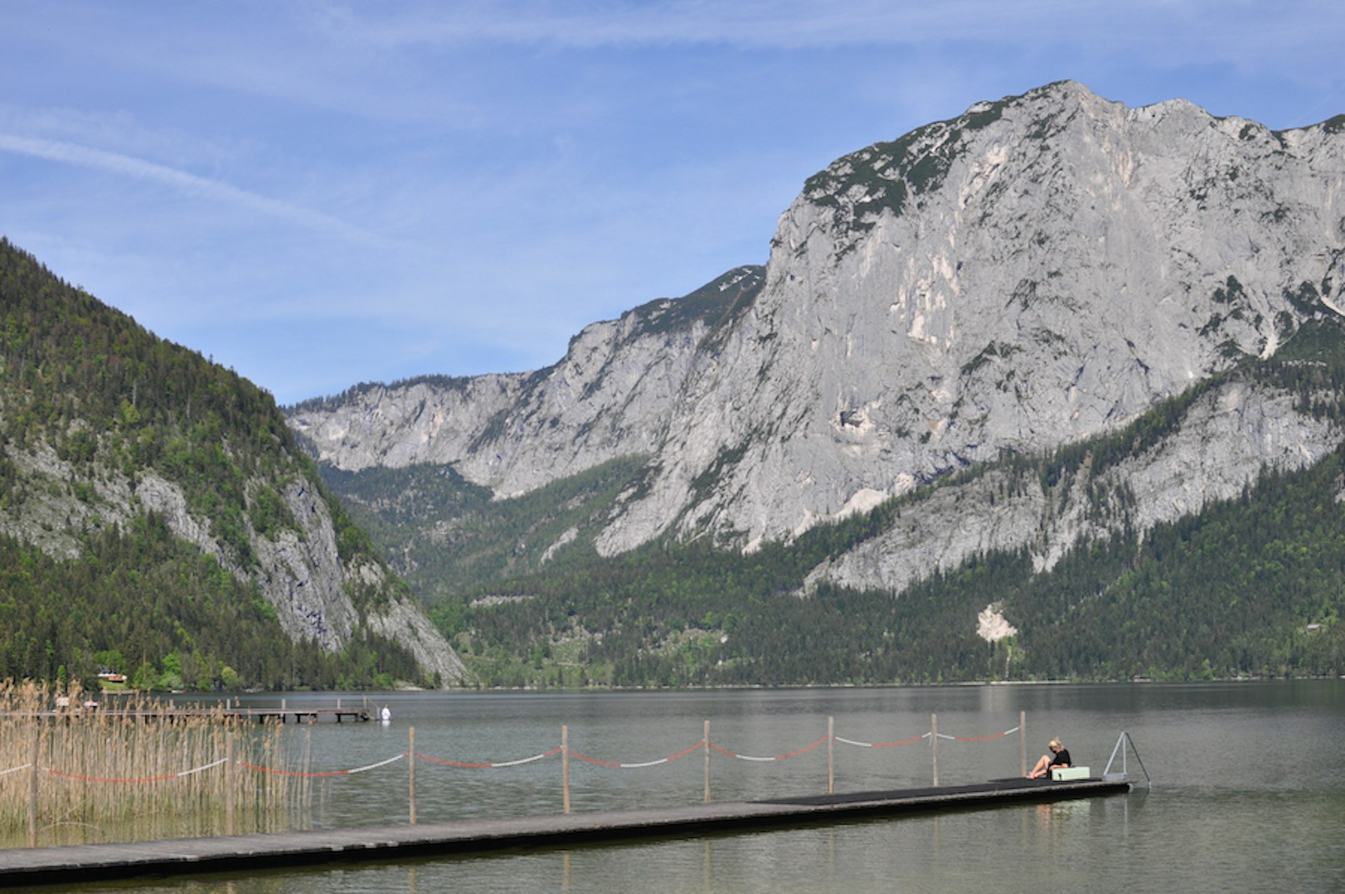 A bather sits on a long dock jutting out into the water with dramatic rocky mountains in the distance at Altausseer see, Upper Austria, Austra
