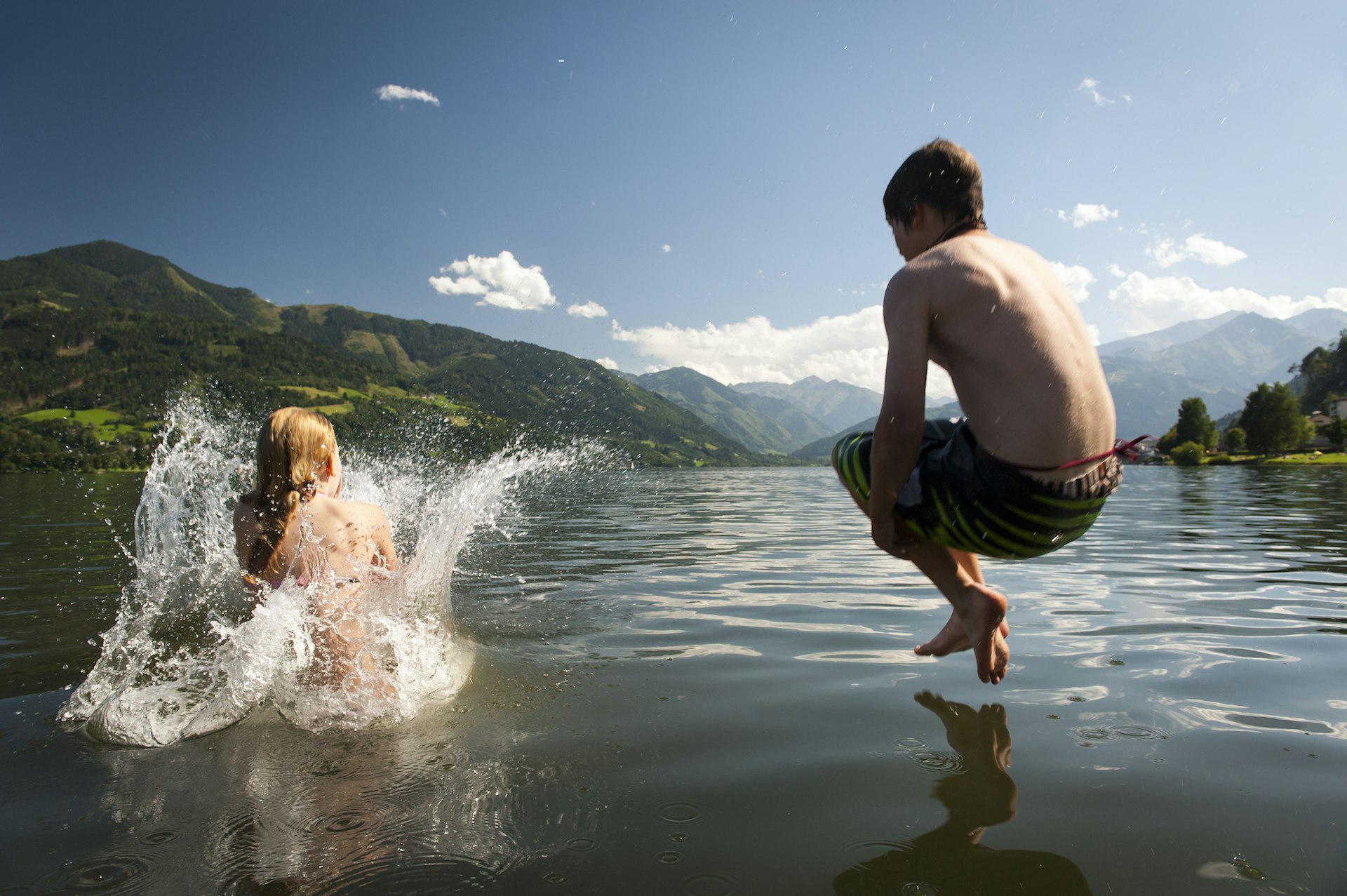 Mountains are seen in the background as a girl and a boy are semi-submerged as they jump into the water of Zell am See, Salzburgerland, Austria