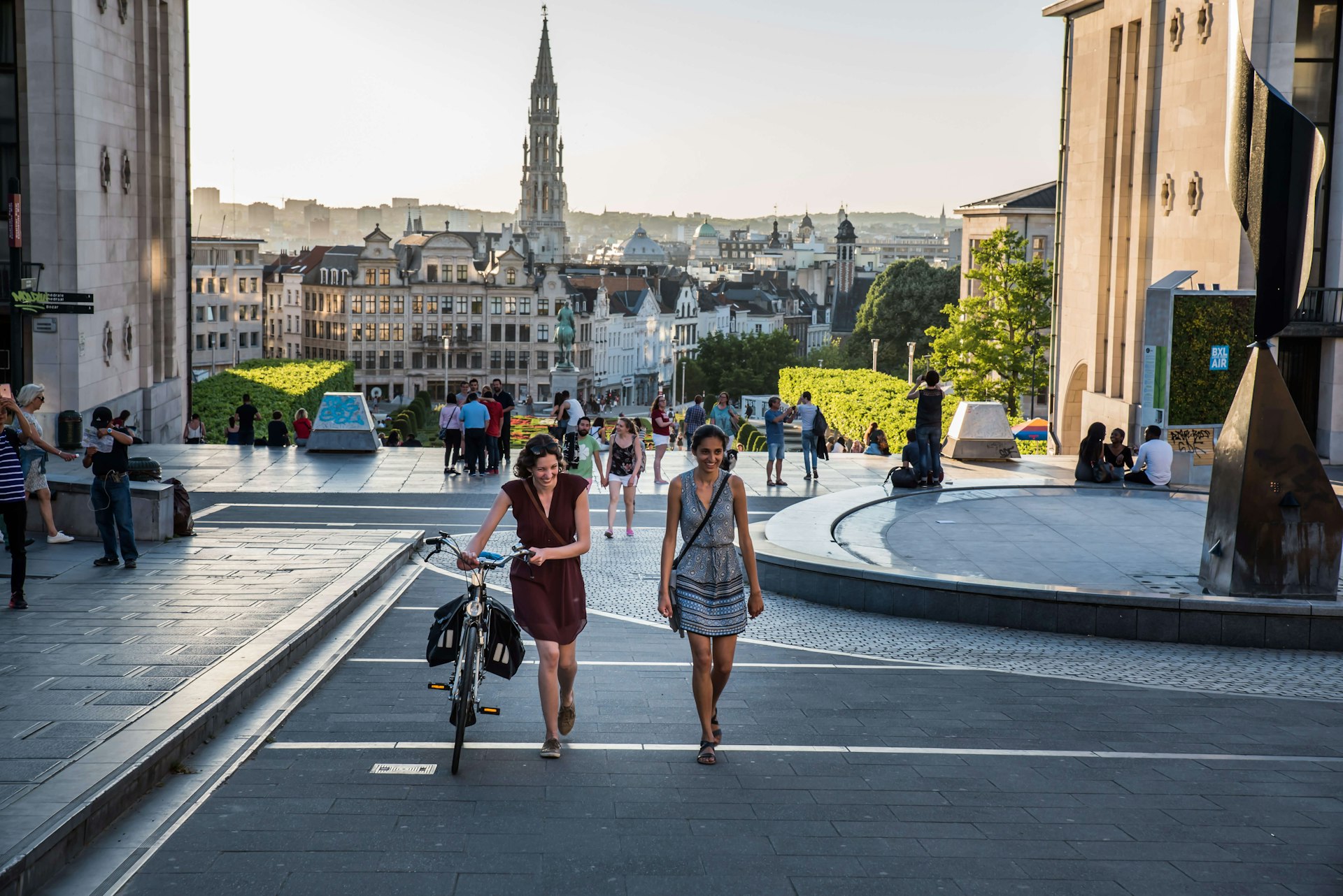 Two young women in summer dresses strolling over sunny streets at Mont des Arts, one pushing a bicycle