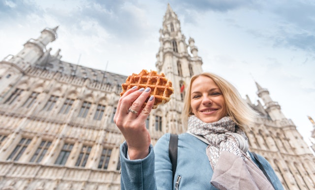 Young woman holds out a traditional Belgian waffle against the background of Grand-Place, Brussels, Belgium