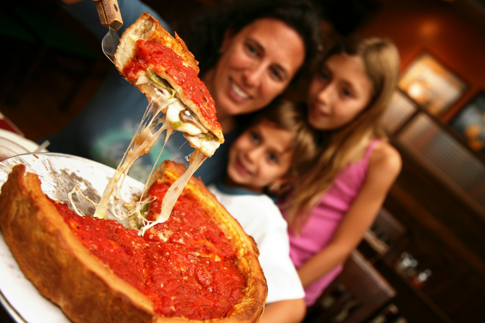 A mom lifts up a slice of Chicago deep dish pizza from a pie with spatula as two young children watch. 
