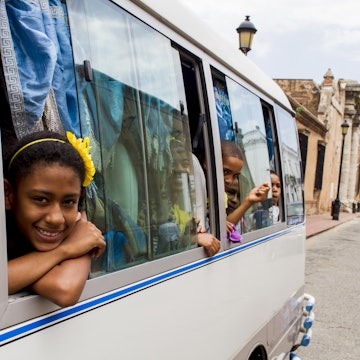 Children peek out of windows of a bus while traveling down a road in Santo Domingo, Dominican Republic.