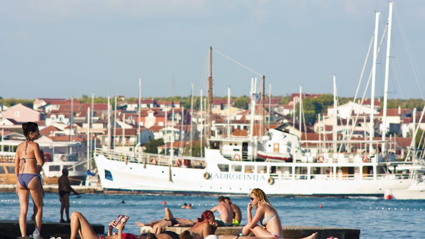 People in swimsuits sunbathe on the pier in Adriatic coast with sailboats, ship and houses in the distance behind them