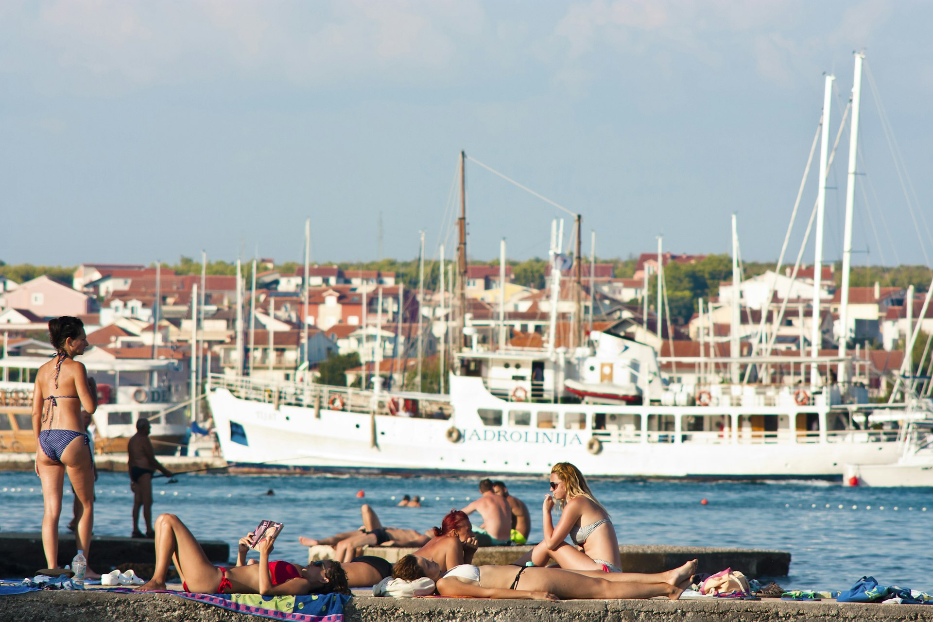 People in swimsuits sunbathe on the pier in Adriatic coast with sailboats, ship and houses in the distance behind them