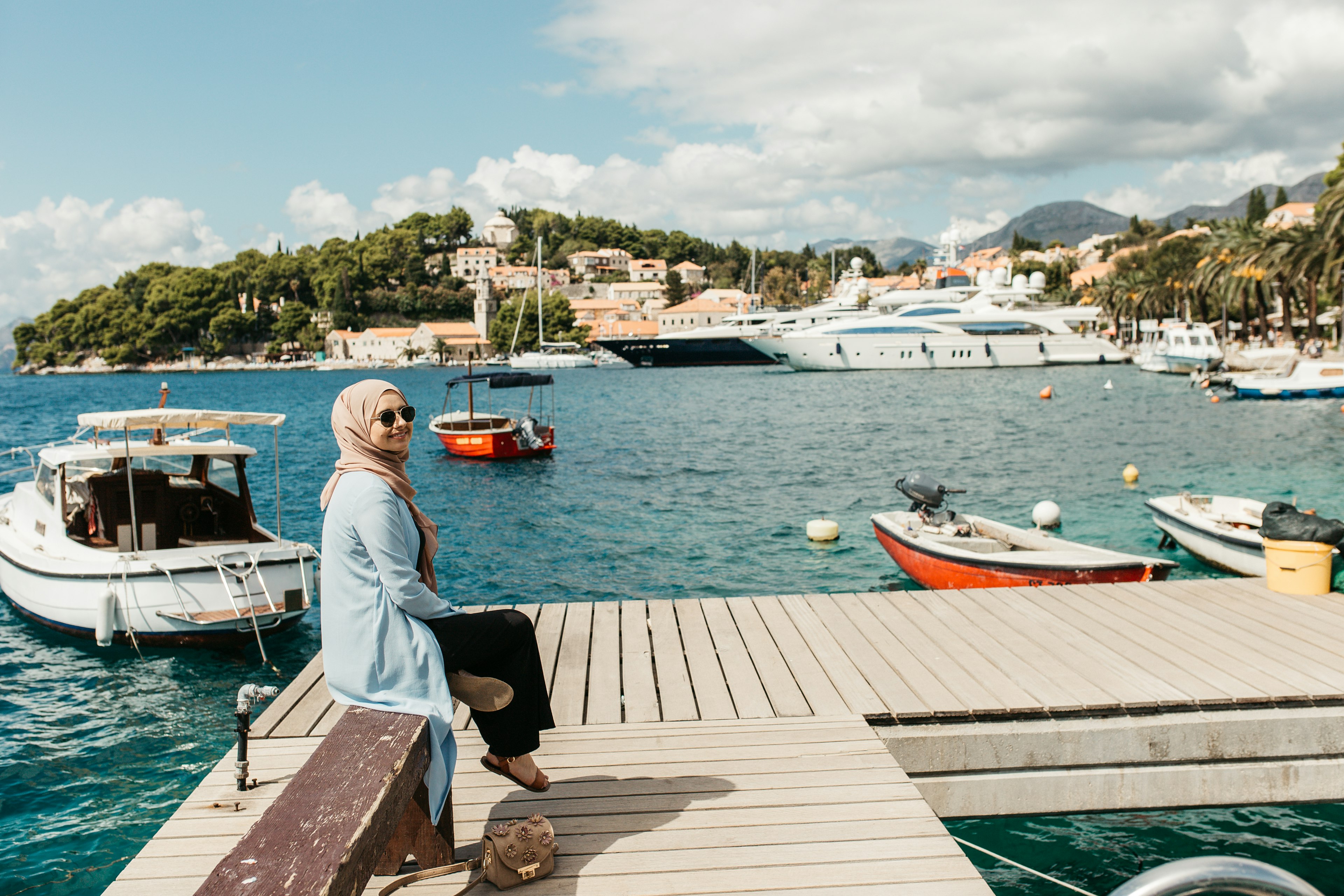 A woman sits on a dock on a sunny day with boats in the water behind her