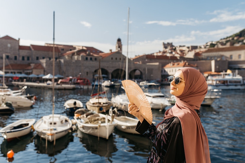 portrait of young European Muslim woman with hijab holding a hand fan and looking at the camera. Sea is in the background. She is happy and relaxed..; Shutterstock ID 1649185399; your: Claire Naylor; gl: 65050; netsuite: Online ed; full: Best time croatia