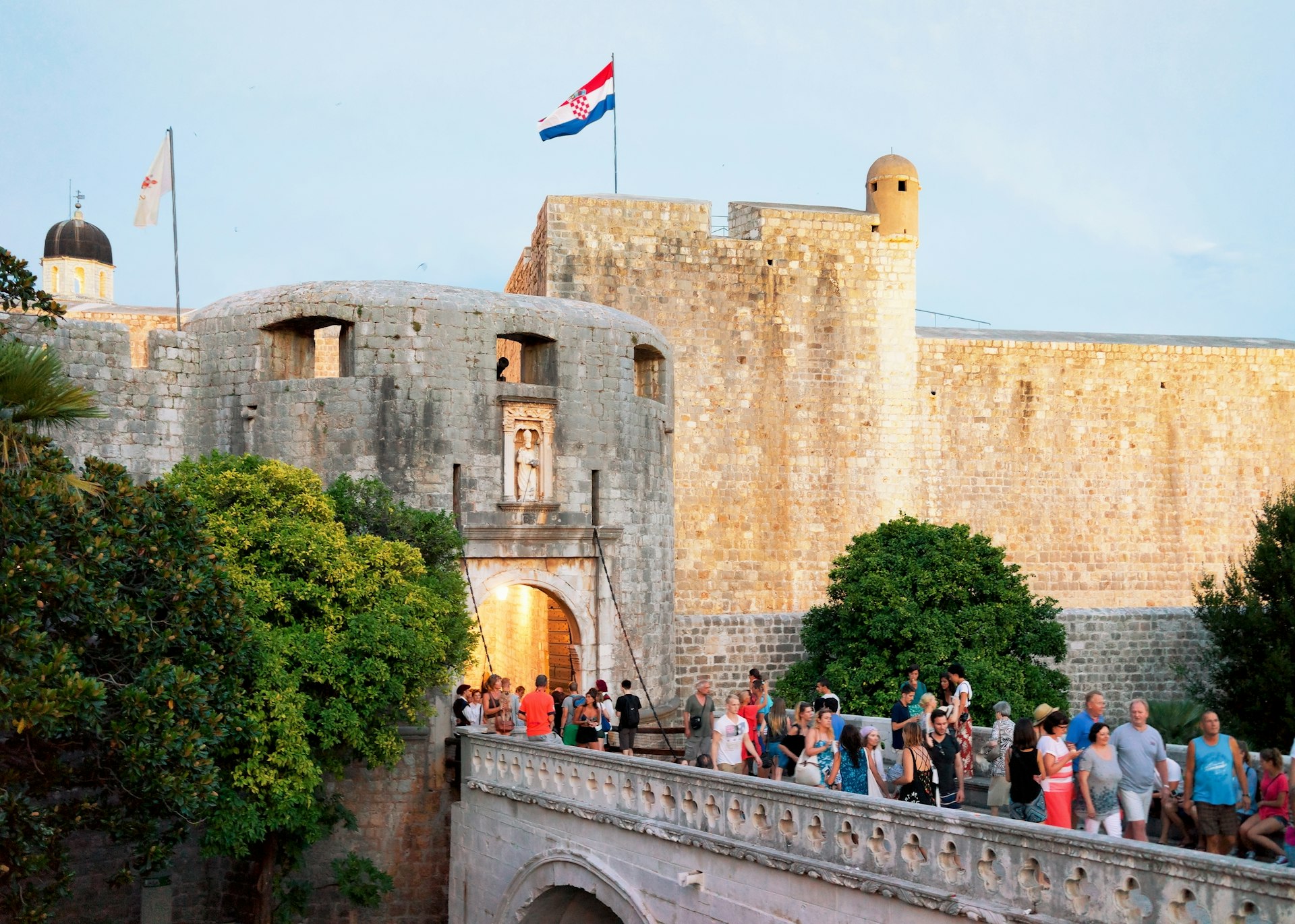 Tourists cross a bridge towards a vast stone gateway in city walls