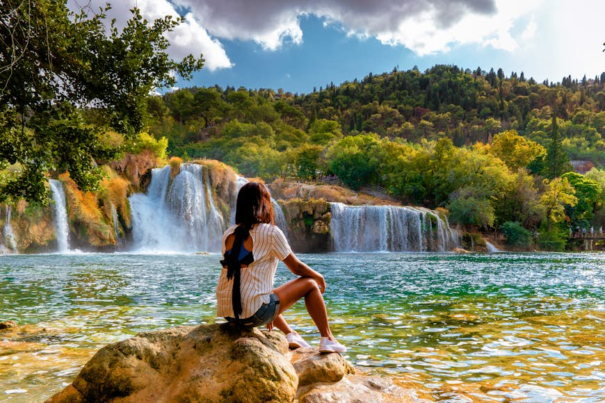 A woman sits on a rock at the edge of a pool being fed by a series of waterfalls in Croatia's Krka national park