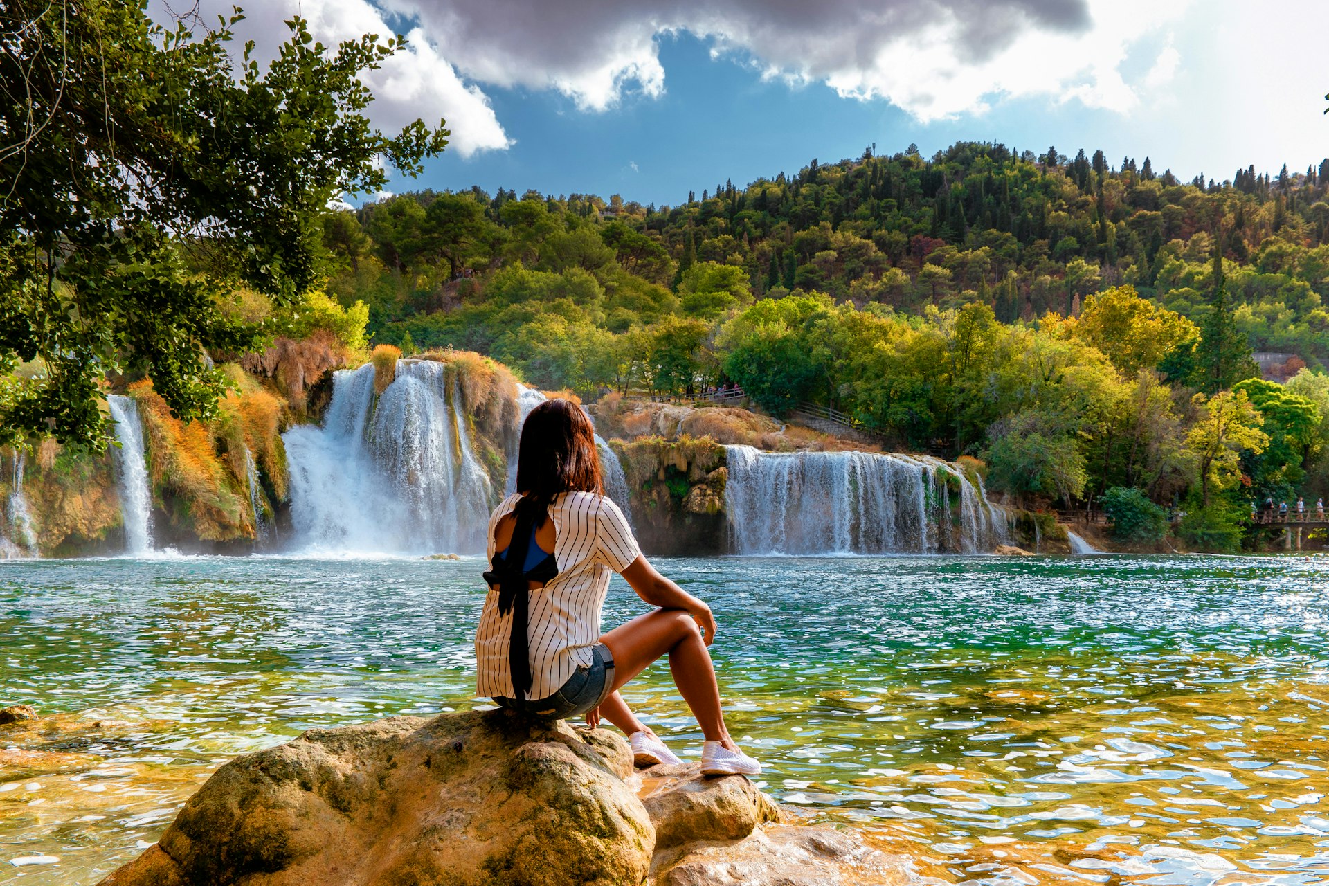 A woman sits on a rock at the edge of a pool being fed by a series of waterfalls in Croatia's Krka national park