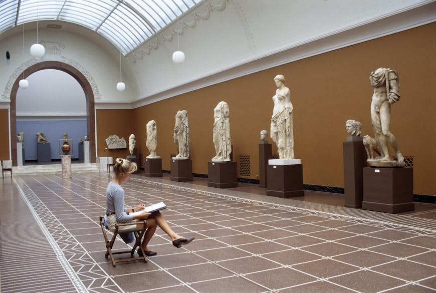 A blonde woman sits in front of classic sculptures while she sketches them in the Glyptotek in Copenhagen