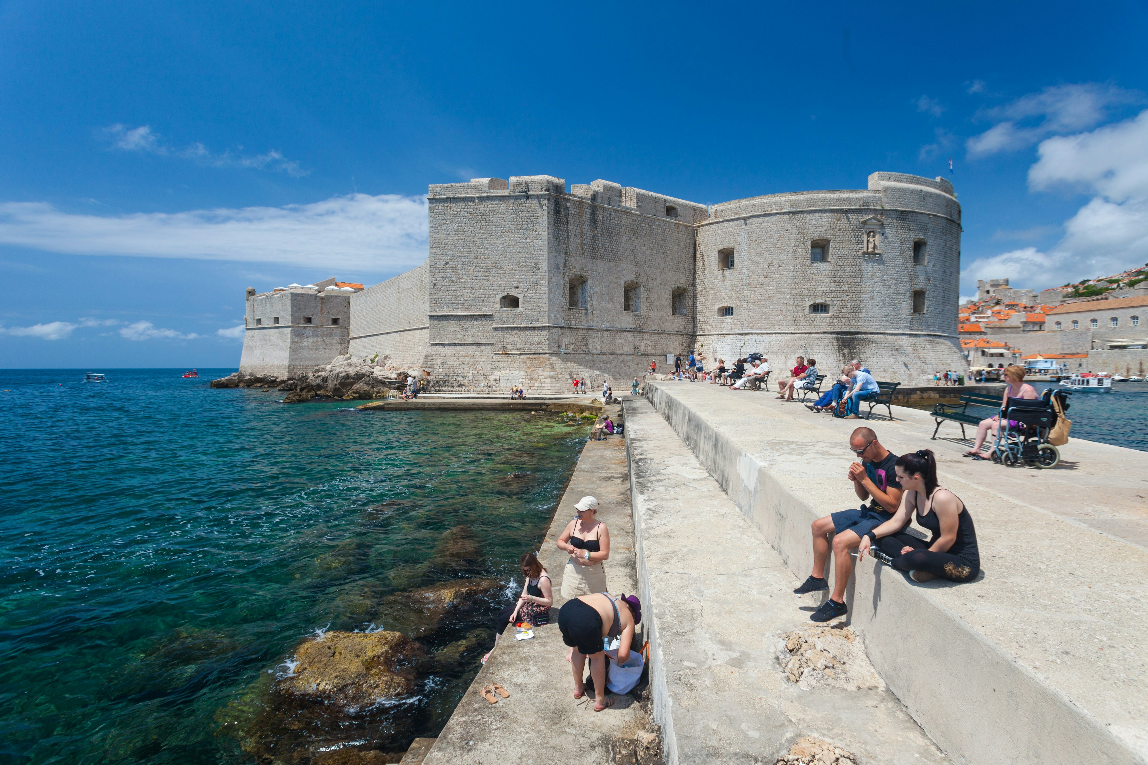 Tourists sitting on bench and stairs on dock in front of the St. John fortress near the old port. Fortress houses the Maritime Museum and the aquarium.