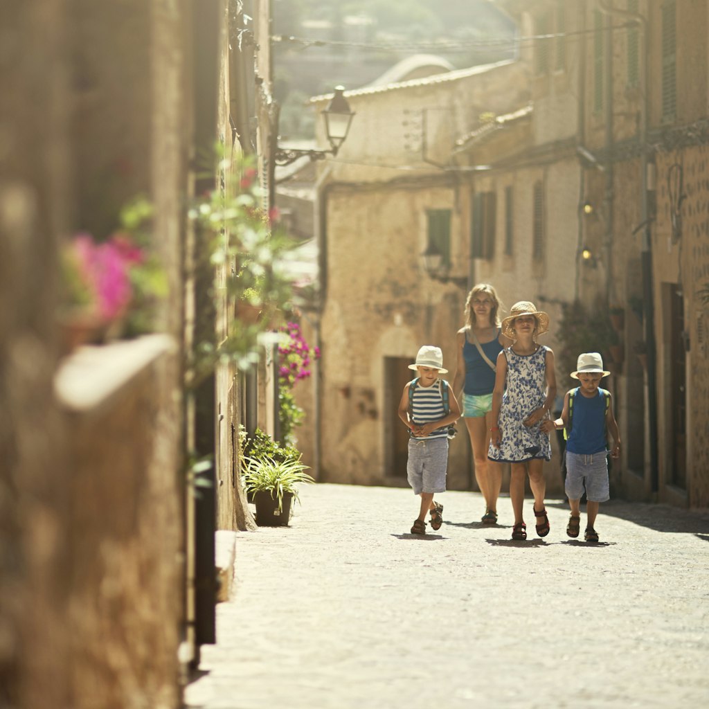 Mother and three kids walking in the beautiful mediterranean town. Little boys are wearing backpacks and hats. Theirs sister is wearing sundress. Sunny summer day. Mallorca, Spain, Valdemossa.