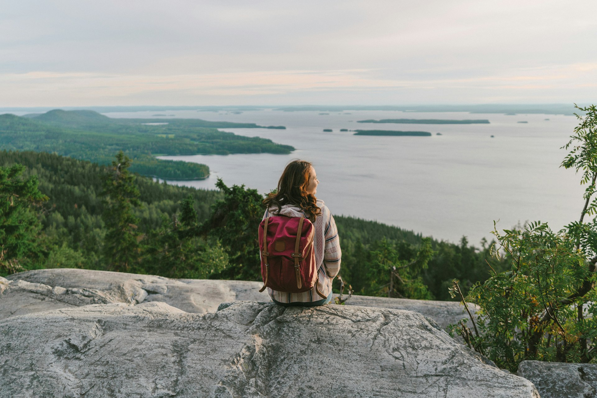 A woman sits on a cliff at the edge of a lake looking out towards forested islets that dot the landscape