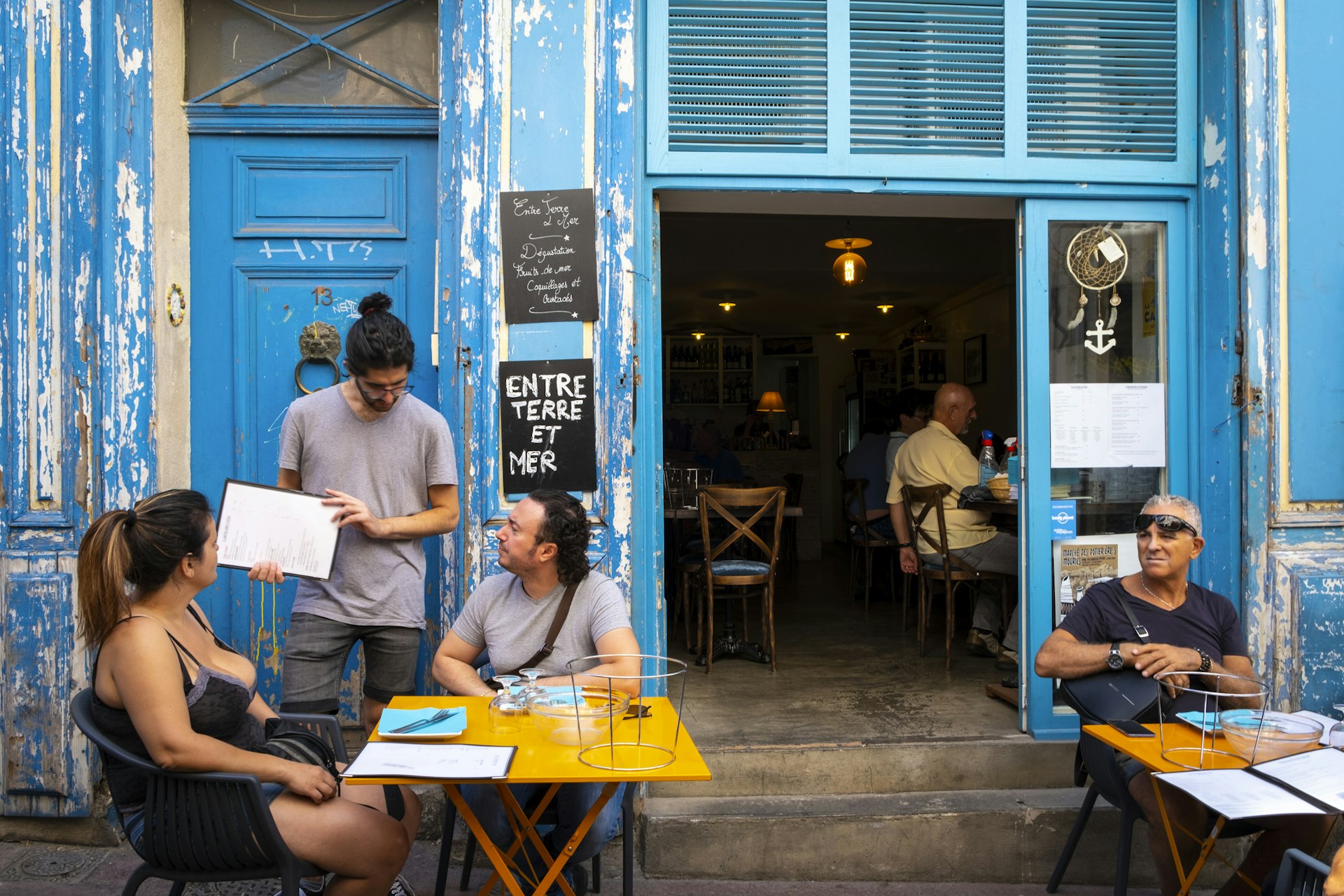 A waiter serves two people sat at a table outside a cafe with a blue facade