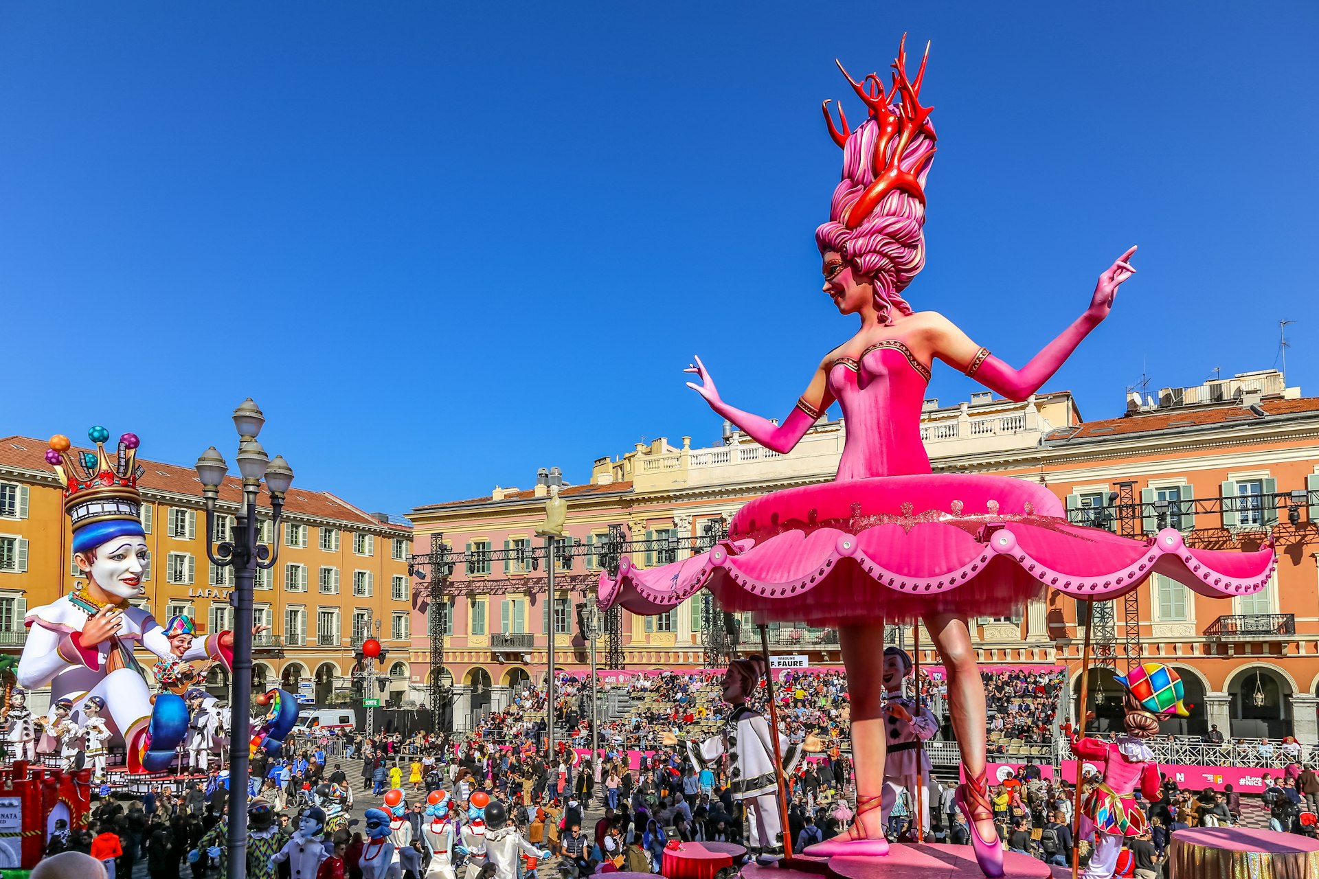 A huge pink ballerina sculpture dominates a carnival float in Nice, France. Crowds of people mingle around the parade
