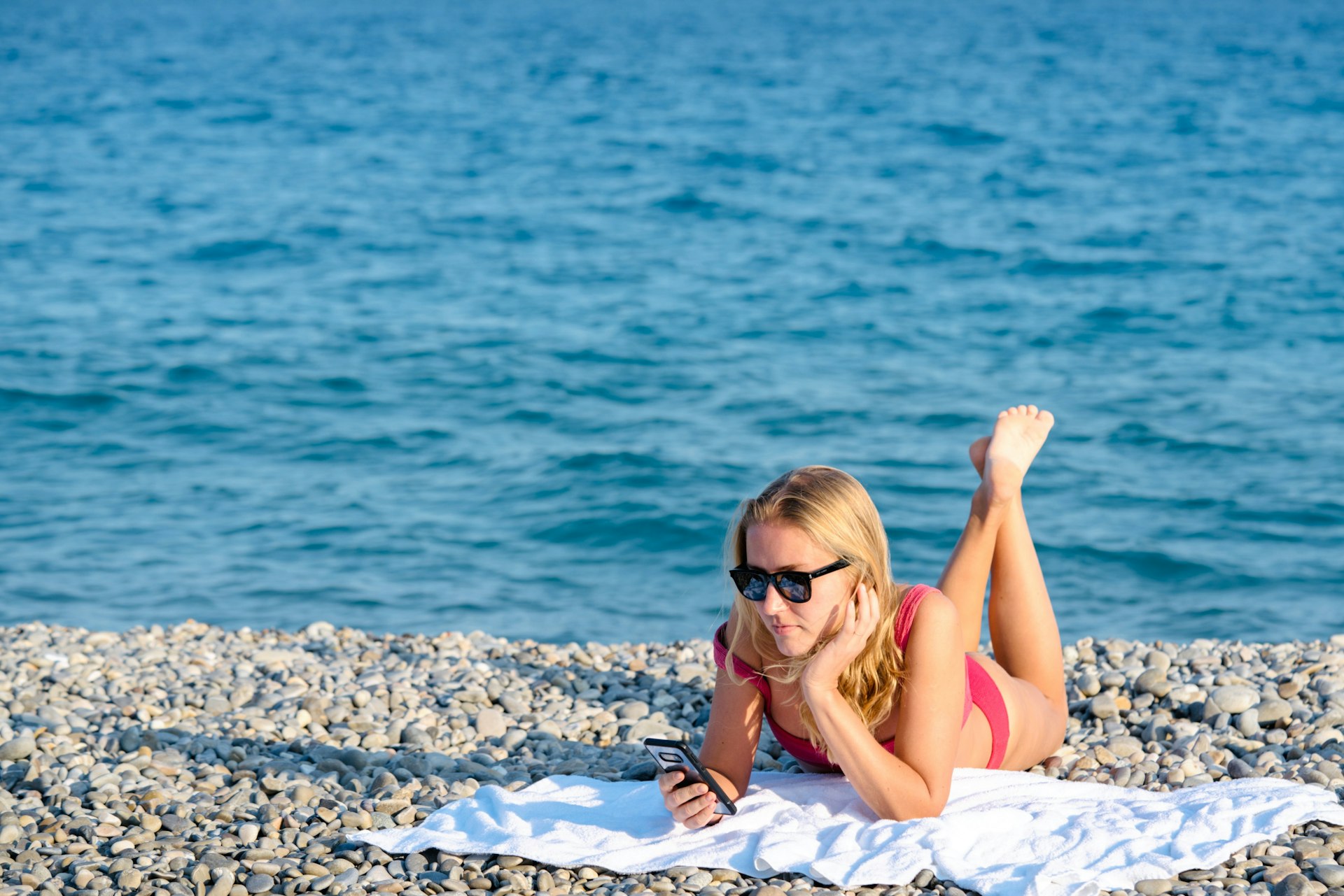 A woman checks her smartphone lying out on a towel on a pebble beach
