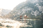 Surface level view of energetic mid adult couple jumping off paddleboard into lake while on summer vacation in Triglav National Park.