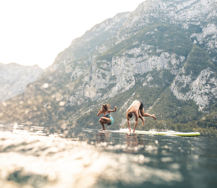 Surface level view of energetic mid adult couple jumping off paddleboard into lake while on summer vacation in Triglav National Park.