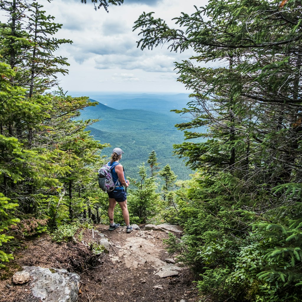A woman is standing at an opening with a view point while climbing a mountain on the Appalachian Trail in Maine.
