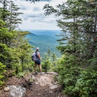 A woman is standing at an opening with a view point while climbing a mountain on the Appalachian Trail in Maine.