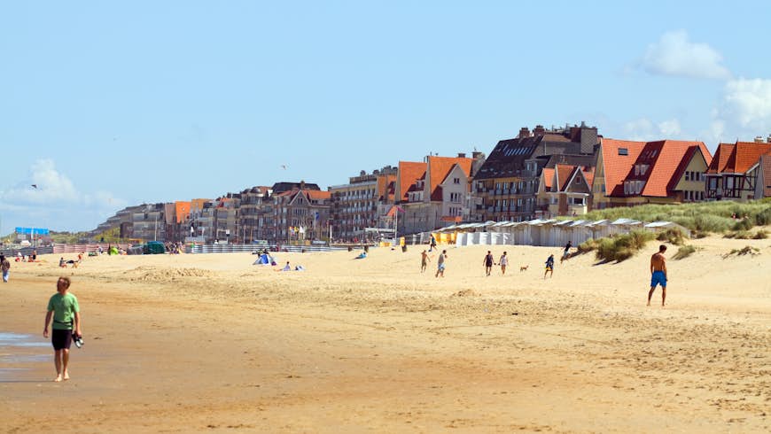 Summer day at the beach in De Haan, on the Belgian North Sea coast, with people walking along beach and a view of a promenade with hotels. 