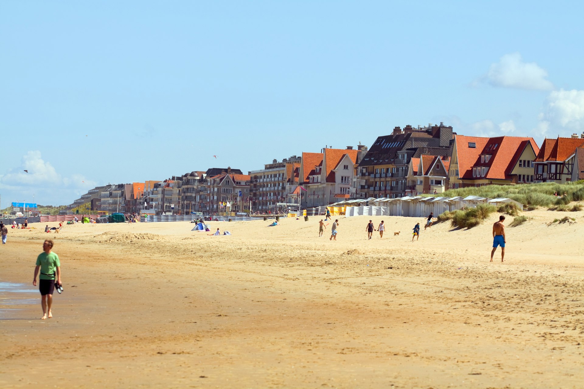 Summer day at the beach in De Haan, on the Belgian North Sea coast, with people walking along beach and a view of a promenade with hotels. 