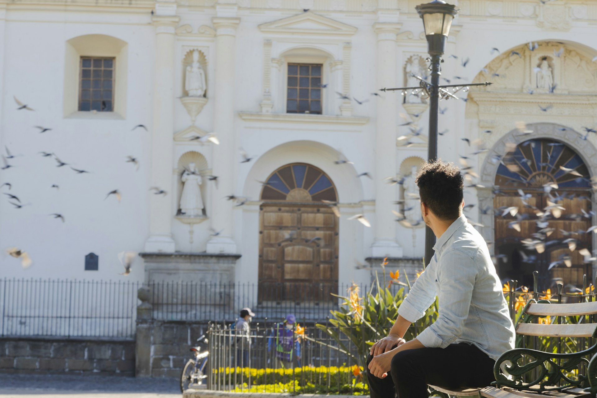 A Hispanic young man sits in front of a cathedral in Antigua, Guatemala, watching pigeons fly over the church
