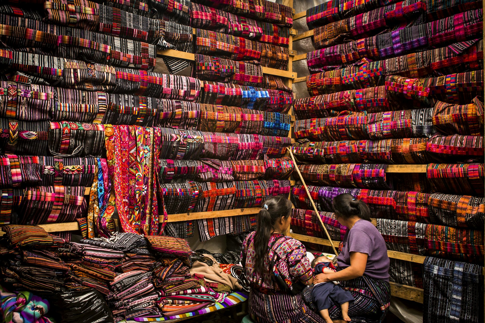 Two women sit in a vendor stall covered in colorful textiles in Chichicastenango, Guatemala