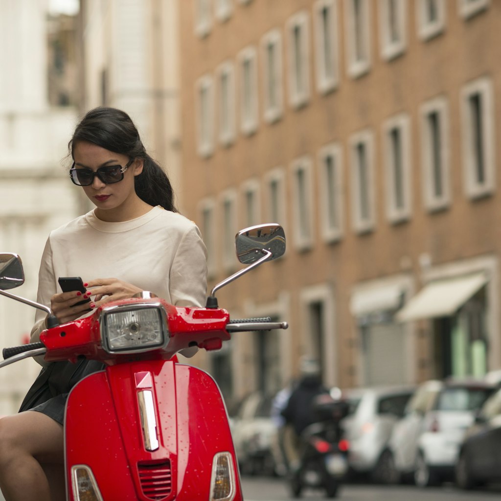 A woman pauses to check her phone on a red moped in Rome