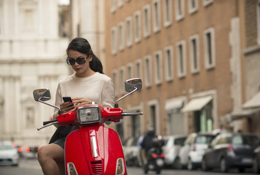 A woman pauses to check her phone on a red moped in Rome