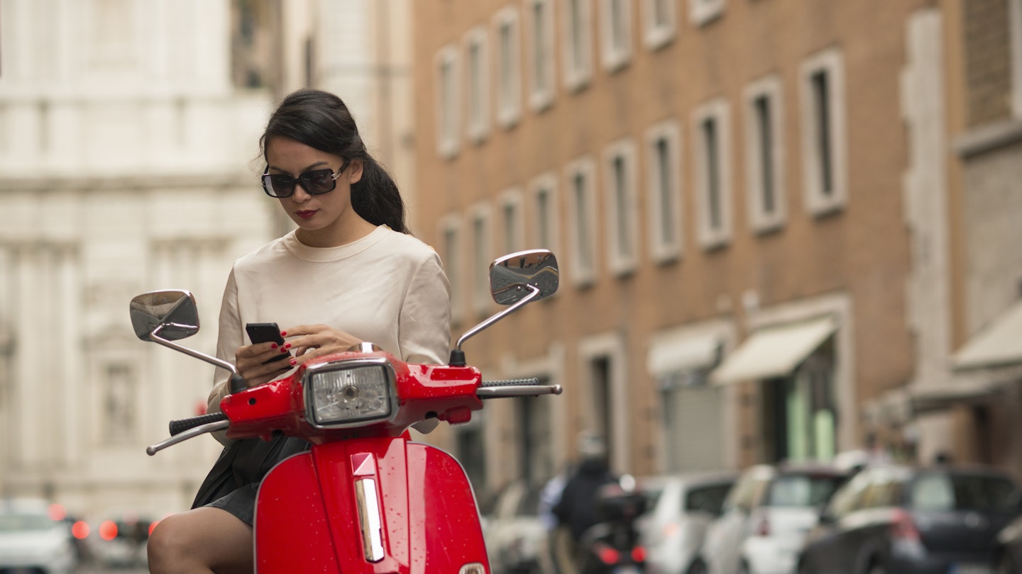 A woman pauses to check her phone on a red moped in Rome