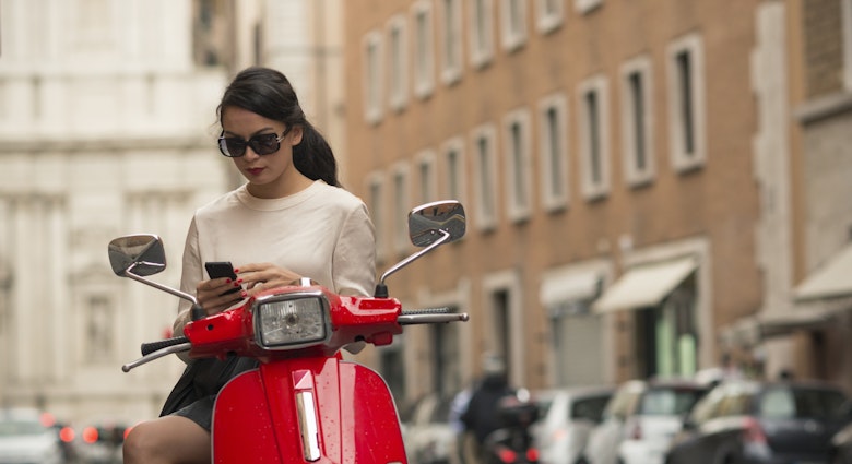 A woman pauses to check her phone on a red moped in Rome