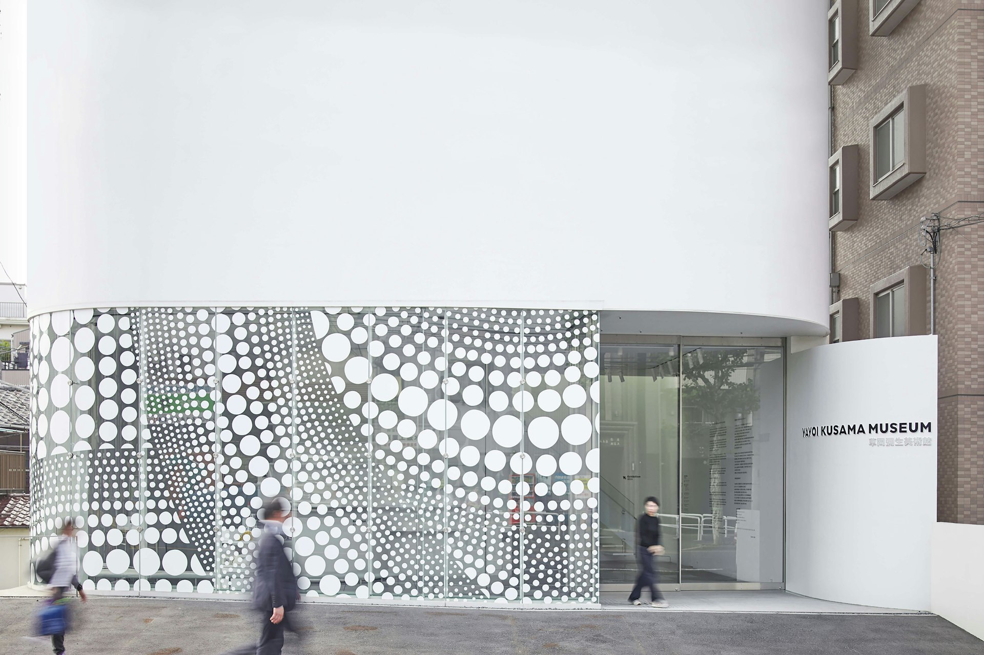 People walking past the Yayoi Kusama Museum in Tokyo, a curved white facade with a polka-dot motif behind glass windows on the ground floor 