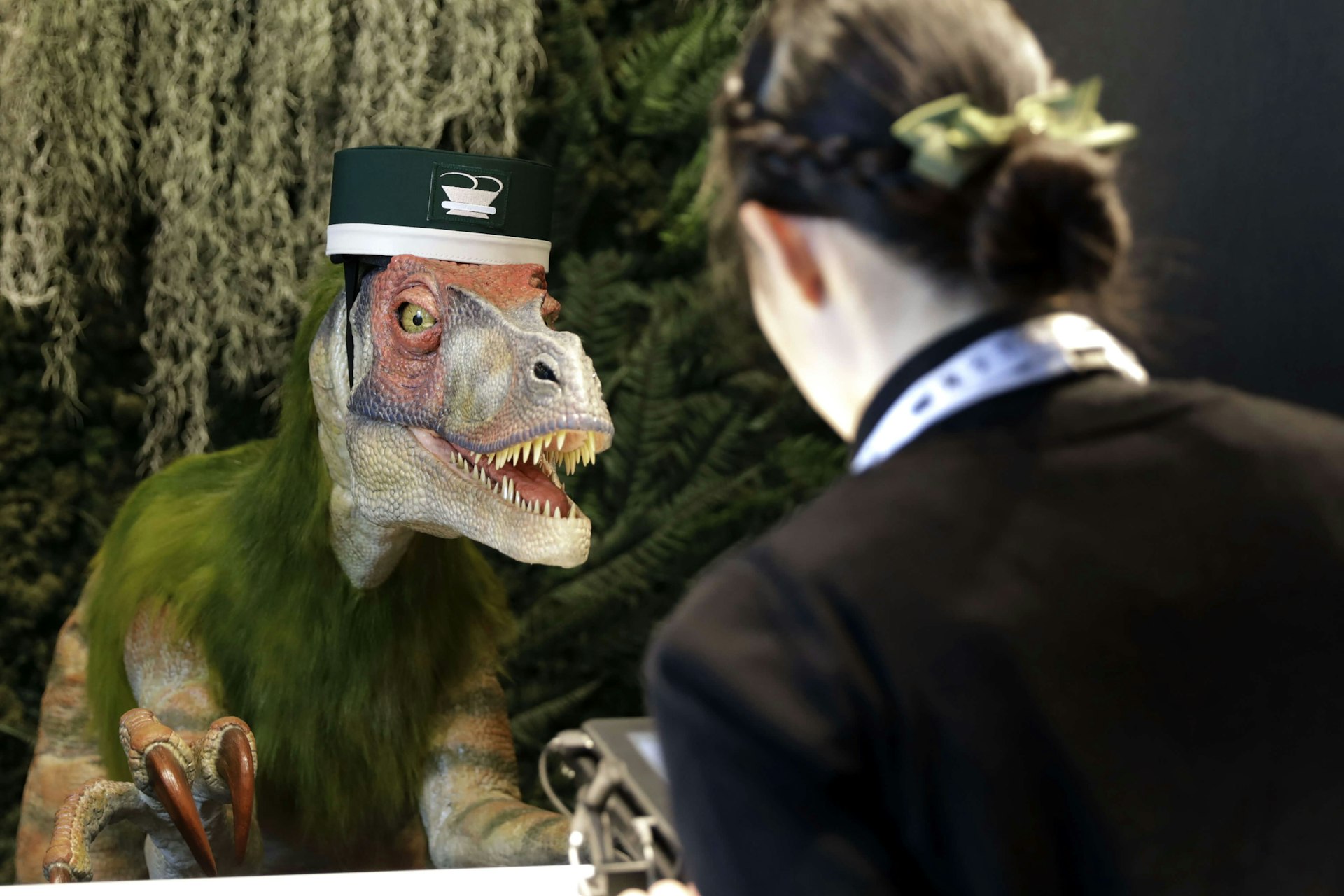 A Kokoro Company Ltd. robotic dinosaur stands at a hotel reception desk as an attendant demonstrates how it works