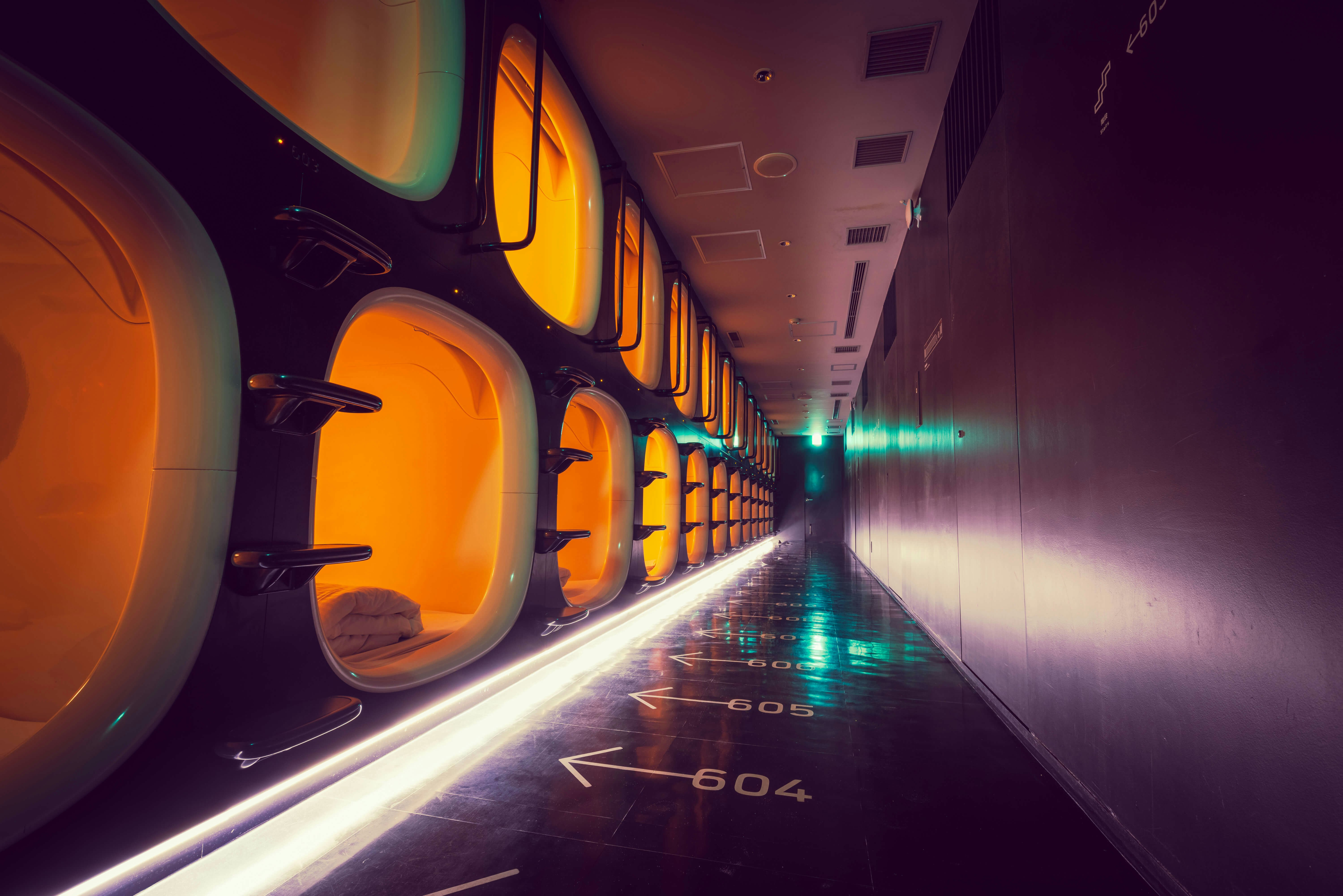 A neon-lit hallway with two levels of sleeping "capsules" running along one wall