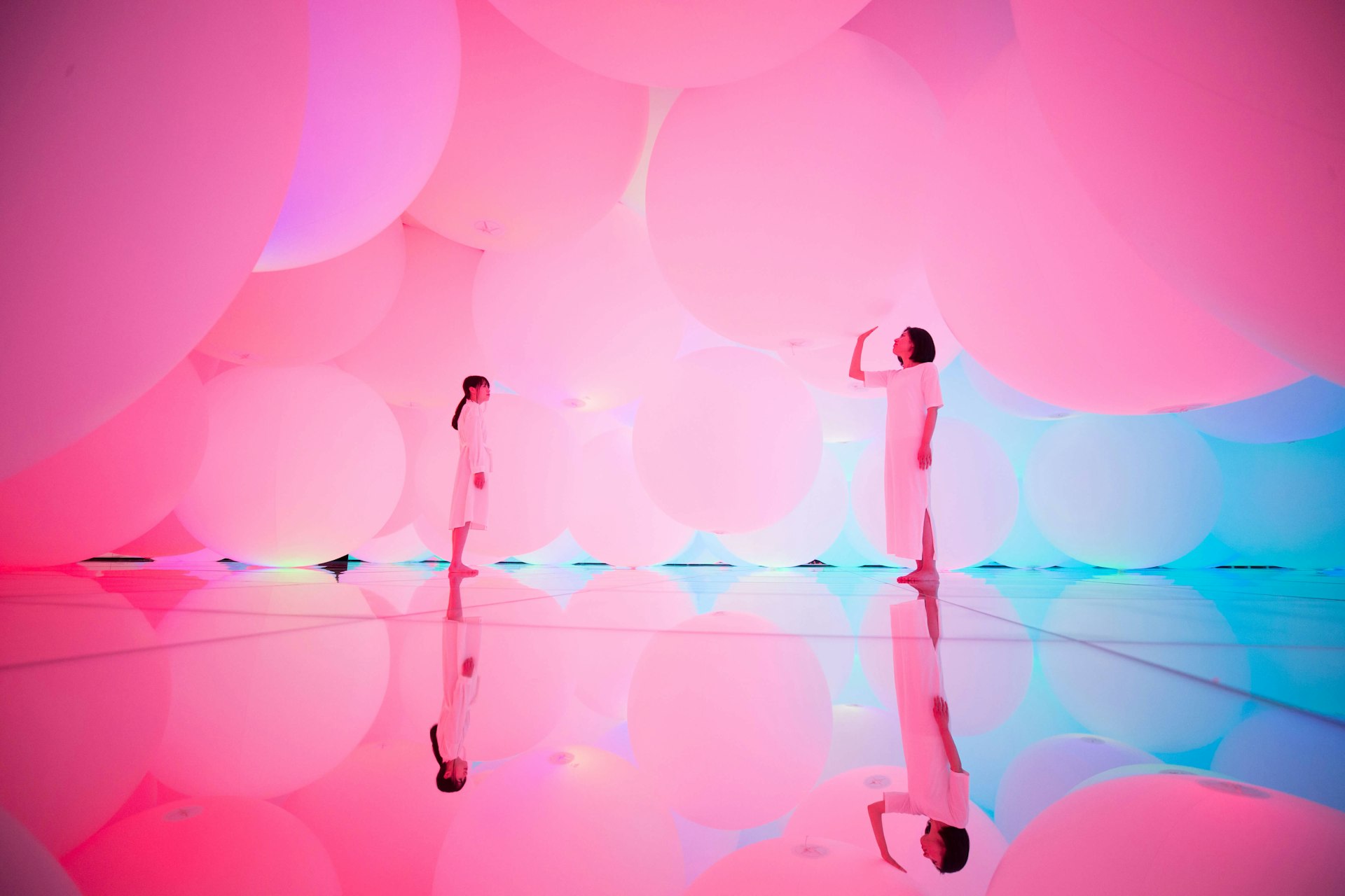 Neon-pink artwork featuring two people in white in a mirrored room filled with hovering globes or balloons