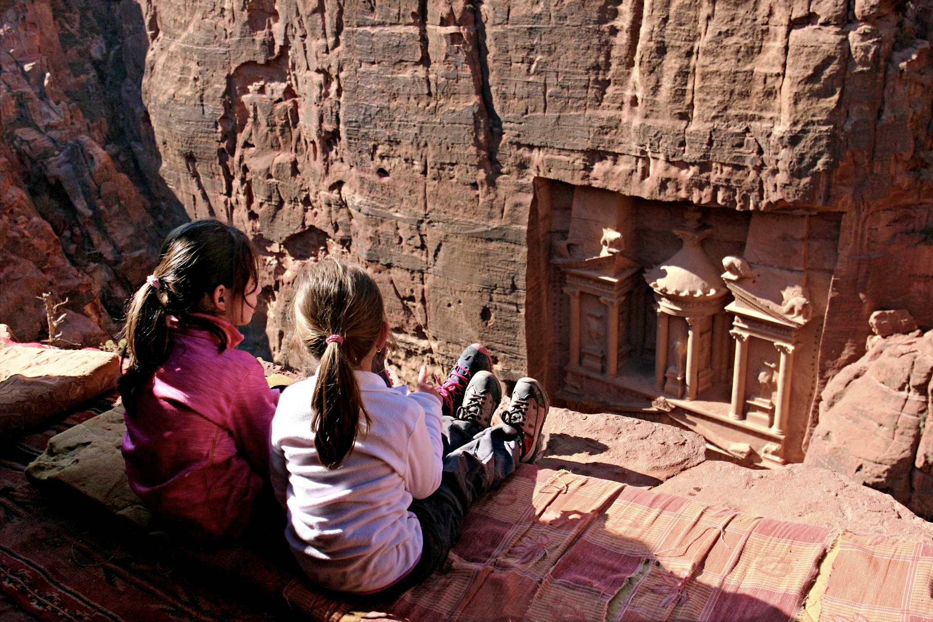 Two young girls sit on a cliff looking downwards into a valley where an ancient city of Petra is carved into the red stone
